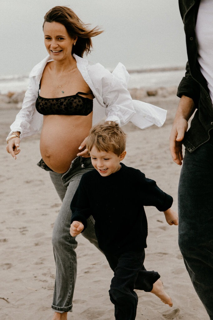 future maman qui court sur la plage avec son fils pendant une séance photo pleine de rire